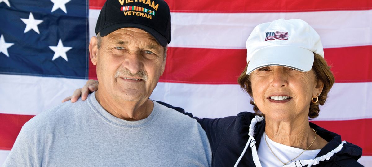 Elderly couple smiling, with the man wearing a Vietnam veterans baseball hat and an American flag in the background